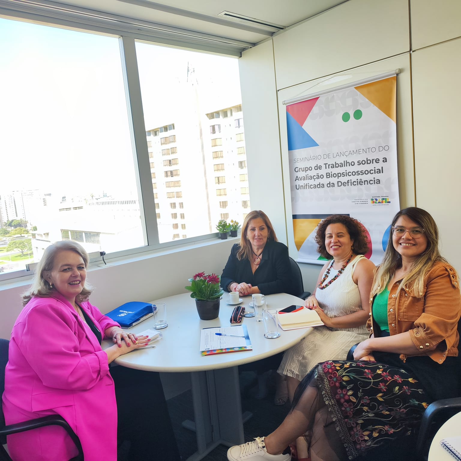 4 mulheres sentadas, sala de reunião, ao fundo banner com texto - Grupo de Trabalho sobre a Avaliação Biopsicossocial Unificada da Deficiência