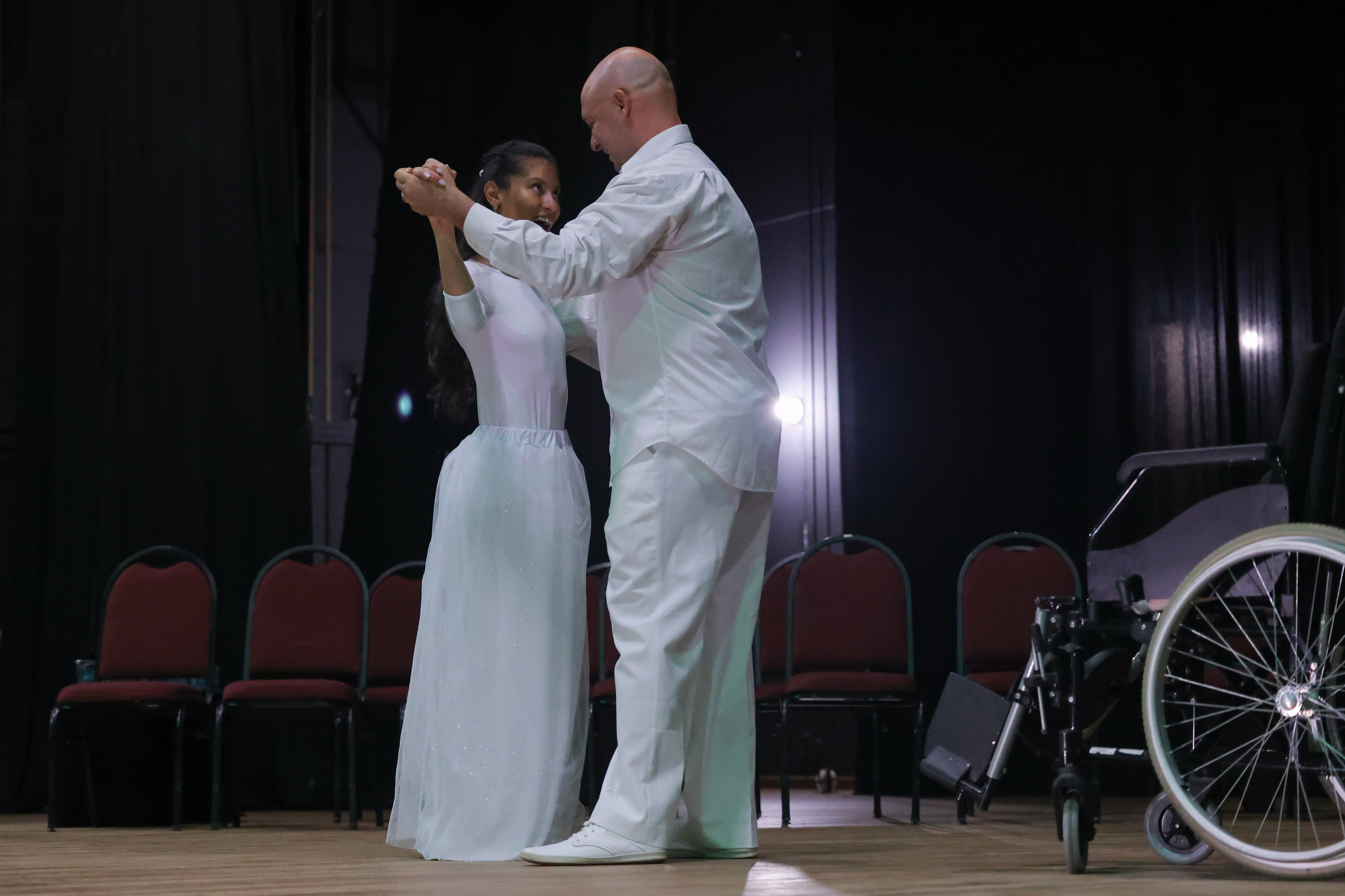 Casal dança em palco vestidos de branco, ao lado uma cadeira de rodas. 