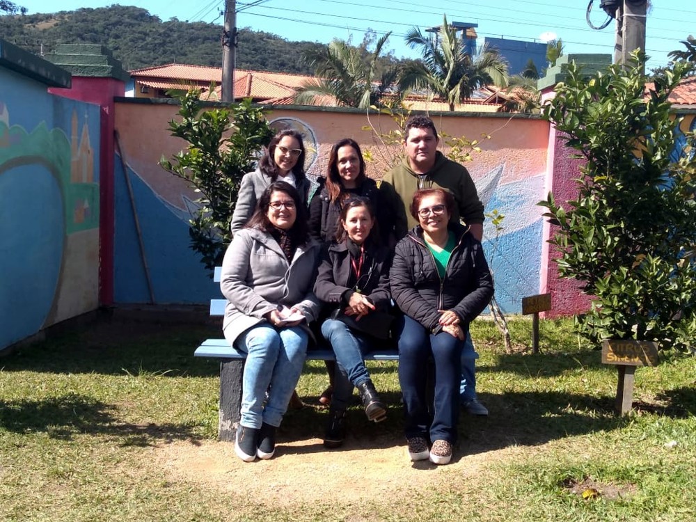 Grupo de seis profissionais, sendo duas profissionais da FCEE e quatro da escola, posam para foto em banco diante de jardim. 