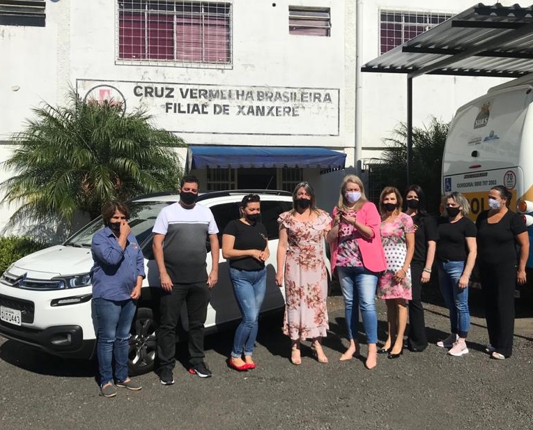 Nove pessoas em pé, ambiente externo, posam para foto, atrás deles um carro branco, ao fundo prédio com letreiro Cruz Vermelha Brasileira Filial Xanxerê