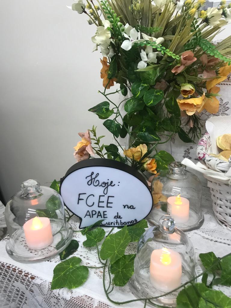 Arranjo de flores em mesa ao lado de arranjos com velas e uma placa escrita com caneta: Hoje: FCEE na APAE de Curitibanos