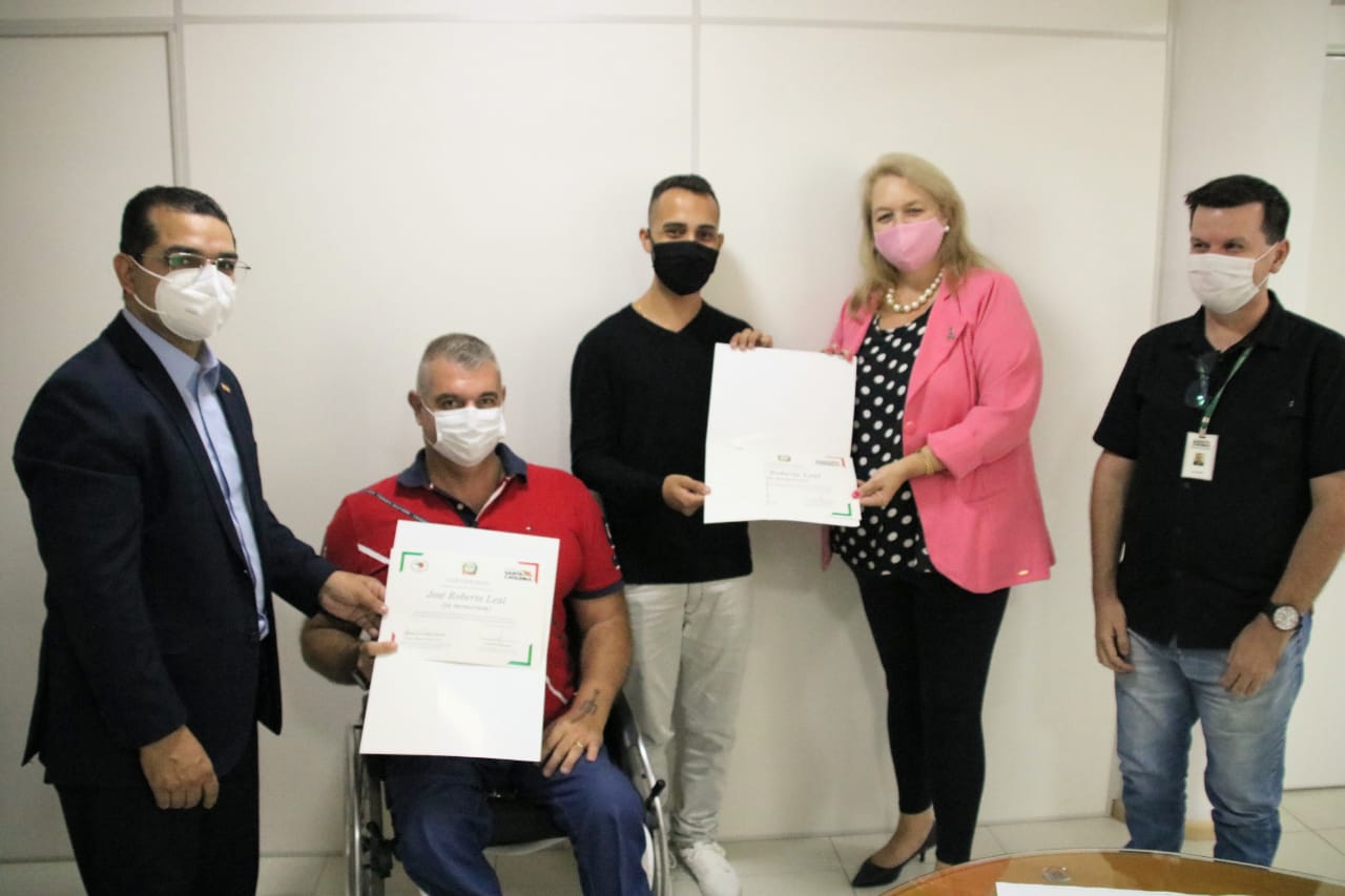 Cinco pessoas posam para foto, um cadeirante, uma mulher e três homens em pé mostram certificados. 