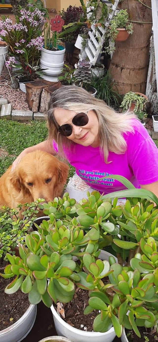 Mulher loira com óculos escuros e camiseta rosa posa para foto ao lado de cachorro bege em meio a jardim com flores. 