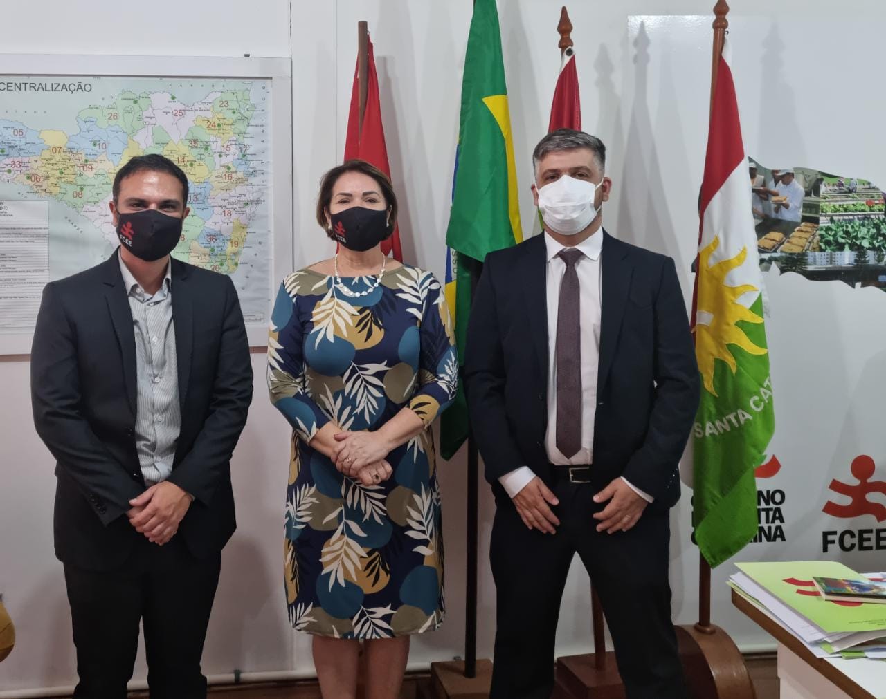 Três pessoas em pé, máscara, ambiente interno, bandeiras ao fundo
