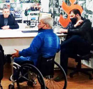 Um homem de cadeiras de rodas em sala de reunião, gesticula com outras pessoas na sala
