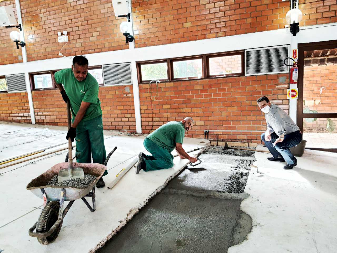 Ambiente interno, chão de concreto, dois homens de uniforme trabalhando no piso, um homem com camisa agachado assistindo. 