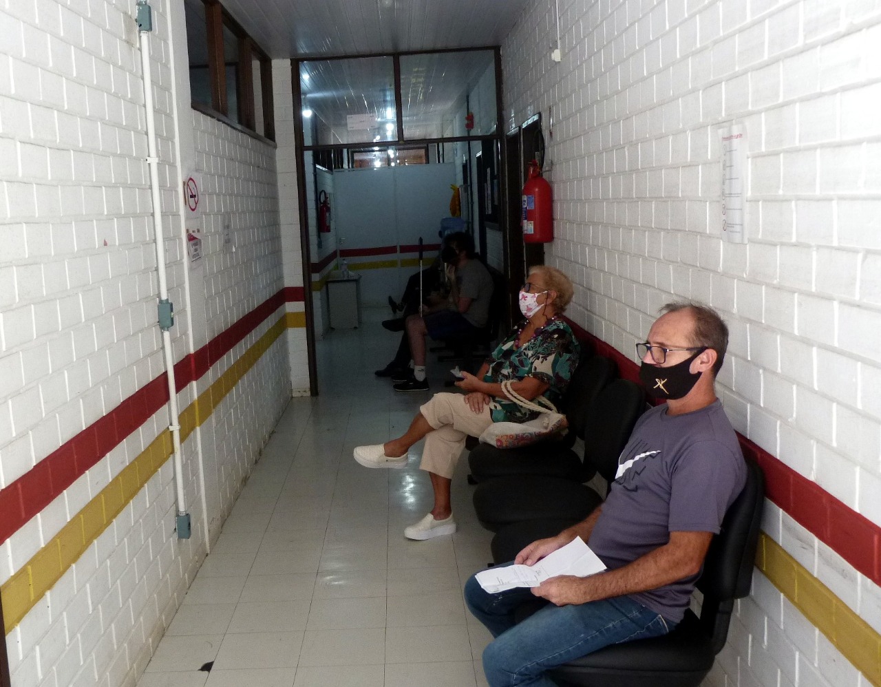 Foto de corredor com longarinas, três pessoas sentadas com distanciamento entre elas.
