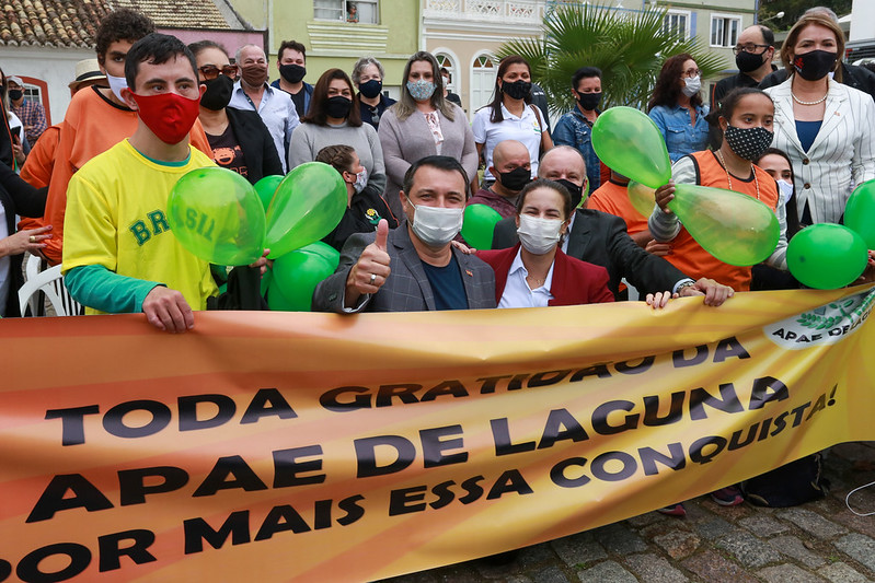 Grupos de pessoas em ambiente externo, seguram faixa laranja com escrita: Toda gratidão da apae de laguna