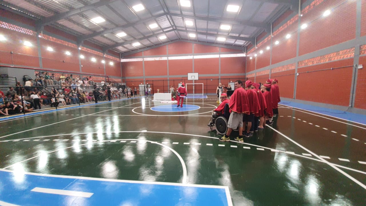 Vista geral do ginásio com pessoas em pé com roupas vermelhas no meio da quadra e ao fundo arquibancada com muitas pessoas sentadas. 