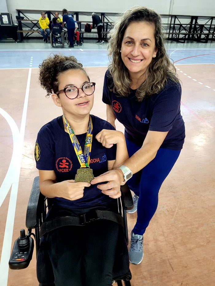 Foto de duas mulheres posando para a câmera, uma menina em uma cadeira de rodas mostra sua  medalha de ouro sorrindo. Ao seu lado, uma mulher está sorrindo de pé. As duas estão em uma quadra esportiva.  