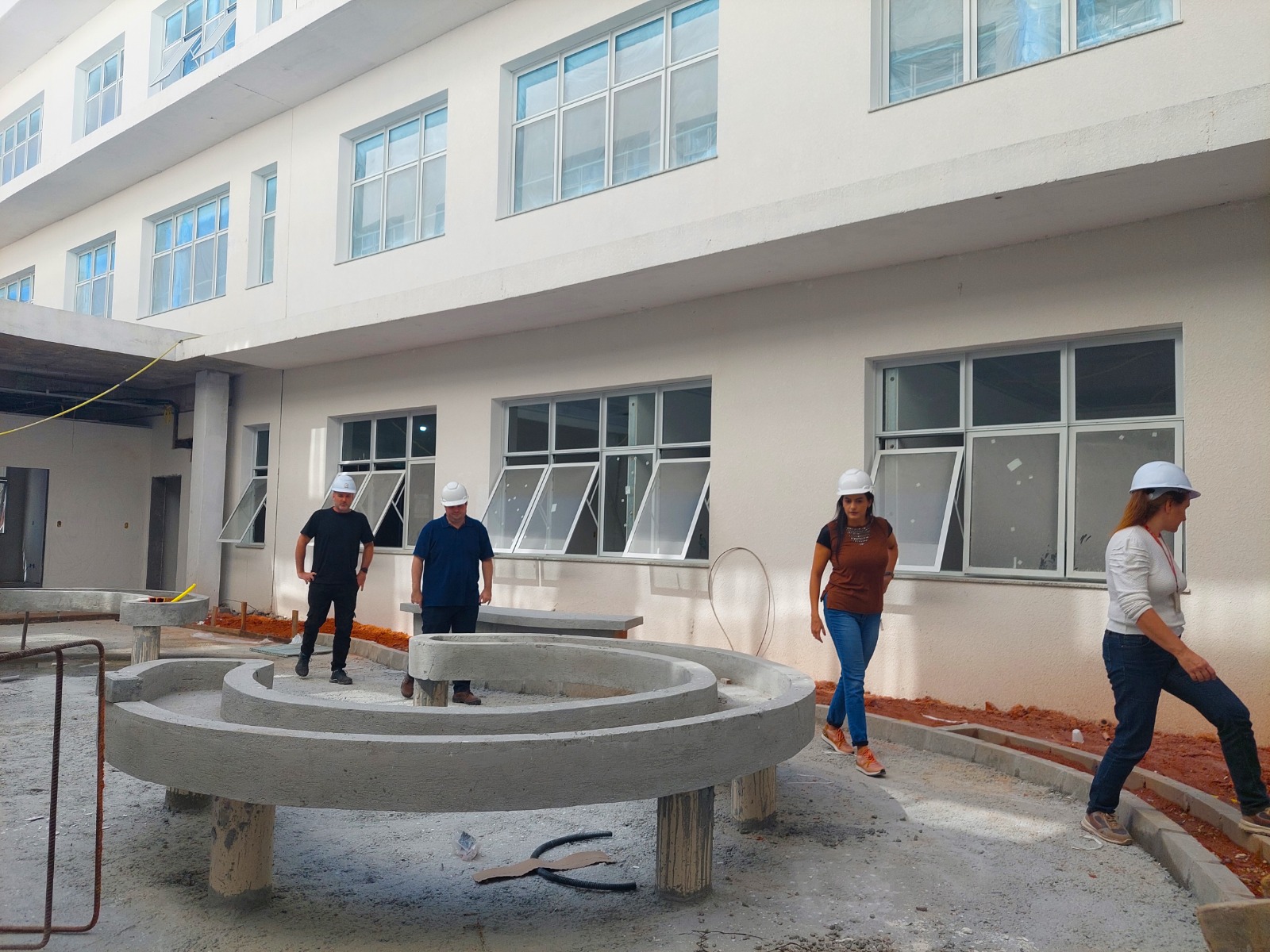 um grupo de quatro pessoas visita as obras em construção do novo prédio da FCEE. Eles estão de capacete branco sobre a cabeça. Passam por uma parte em concreto do futuro jardim interno