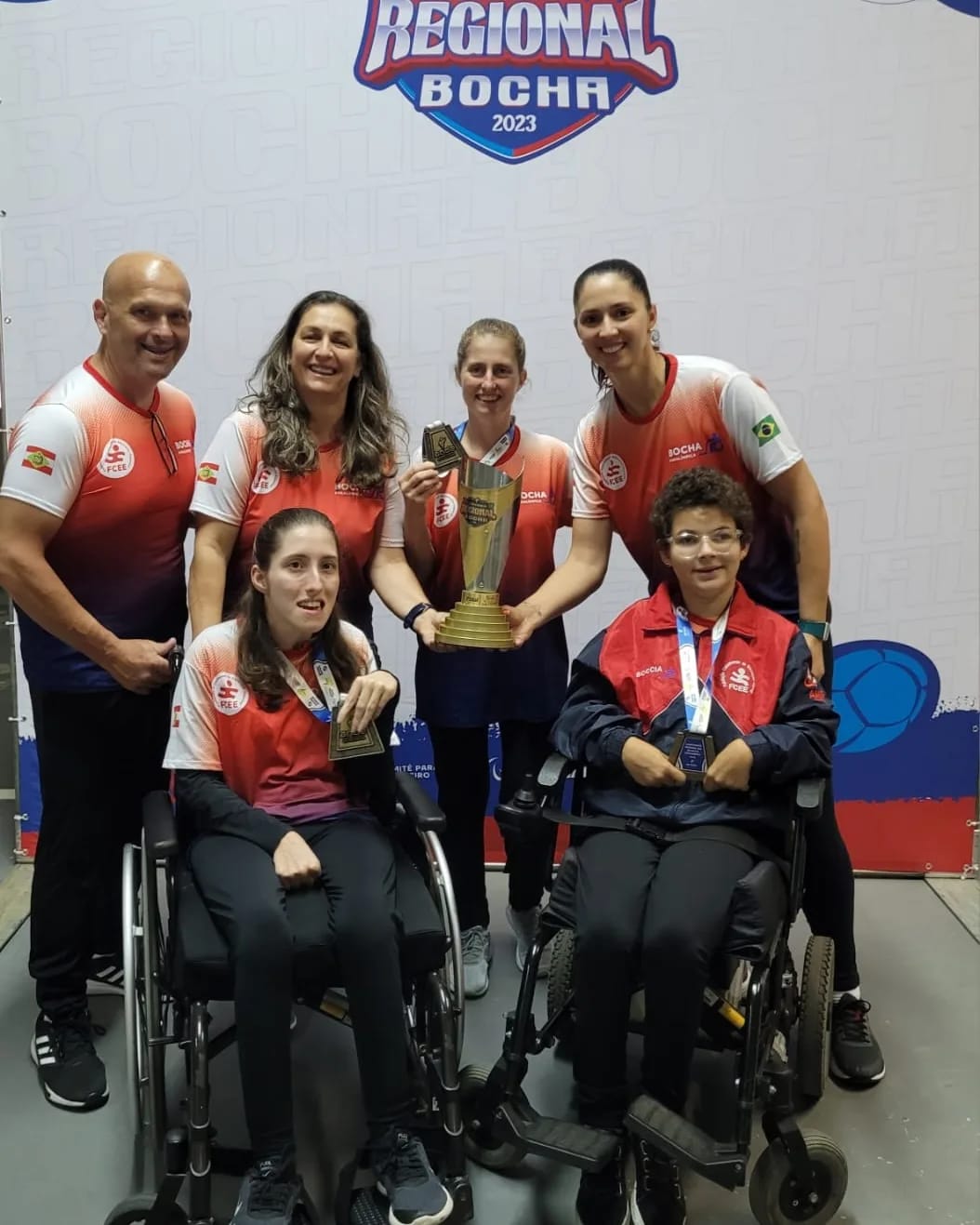 4 pessoas em pé e 2 meninas em cadeiras de rodas posam para foto com medalhas