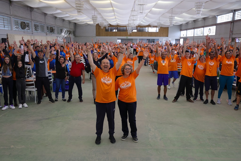 Em um salão, um grupo de atletas e participantes, a maioria trajando camisa laranja, levantam as mãos para o alto, saudando a abertura do Festival Paradesportivos da Apae. A frente o governador Jorgunho Melho e  a presidente da Feapaes-SC, Alice Kuerten