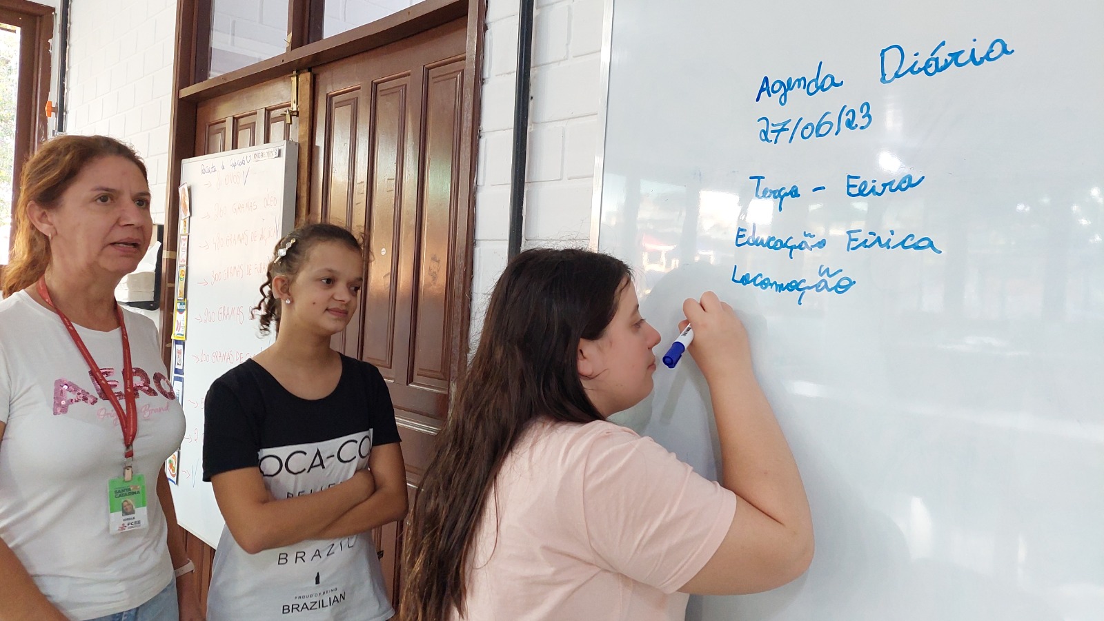 Uma aluna escreve em um quadro branco em sala de aula sendo observada pela professora e uma colega de classe