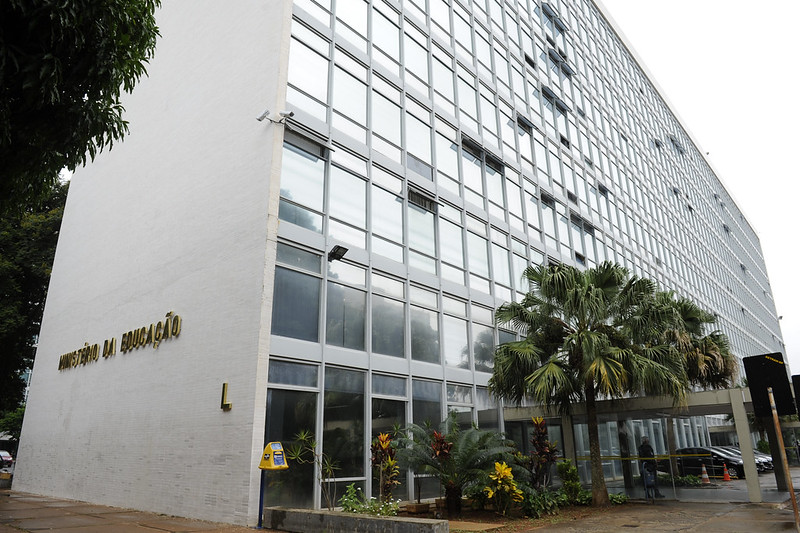 A imagem mostra a fachada do prédio do Ministério da Educação em Brasília. O prédio tem janelas de vidros ante reflexo. Na frente tem uma palmeira e na lateral do prédio um letreiro, onde está escrito Ministério da Educação