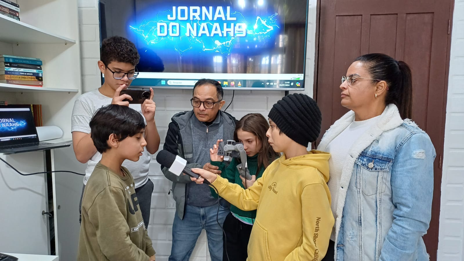 Seis pessoas em uma sala. São quatro adolescentes acompanhados de dois professores, que são um homem e uma mulher. Na frente da foto um aluno moreno, com moleton bege é entrevistado por um outro garoto moreno com moleton amarelo e toca na cabeça ele segura um microfone de repórter. O professor, um homem morenos de moleton cinza observa. Do lado esquerdo um adolescente , moreno, de óculos, filma a cena com um celular. Ao lado do professor uma garoto branca também filma a cena. Ao lado direito uma professora, morena, de cabelo liso e óculos, trajando um moleton jeans observa a cena. Ao fundo uma tv ligada mostra a frase: Jornal do Naahs