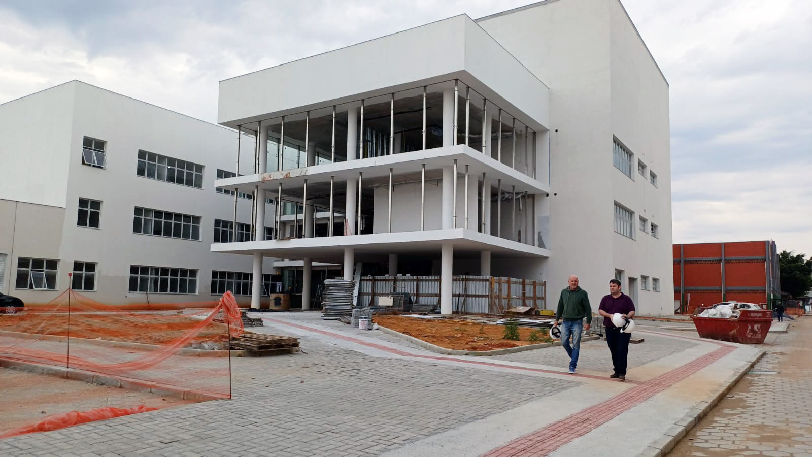 Vista frontal do prédio em construção, 3 pavimentos em cimento, 2 pessoas caminham na calçada. 