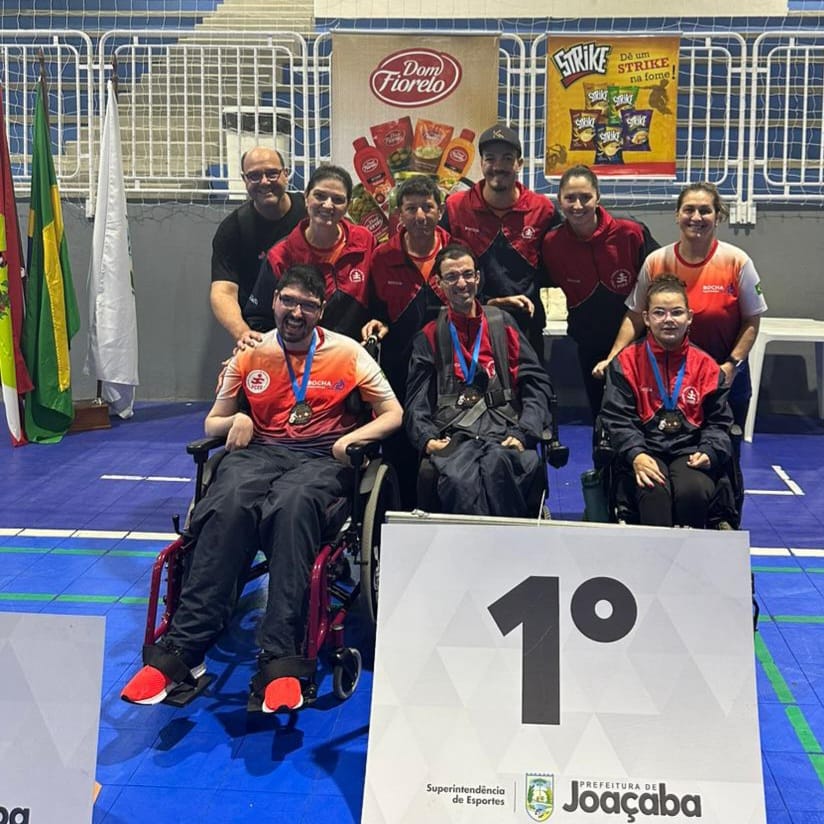 Um grupo de nove pessoas posa para uma foto no pódio de uma competição. A frente três atletas cadeirantes estão com medalhas no peito. Estão de blusa vermelha e calça preta, que fazem parte do uniforme. Atrás deles está a equipe de professores e auxiliares. A frente, em primeiro plano  há número 1, em alusão ao pódio de primeiro lugar. Atrás deles, na parede do ginásio, há cartazes de várias empresas patrocinadores