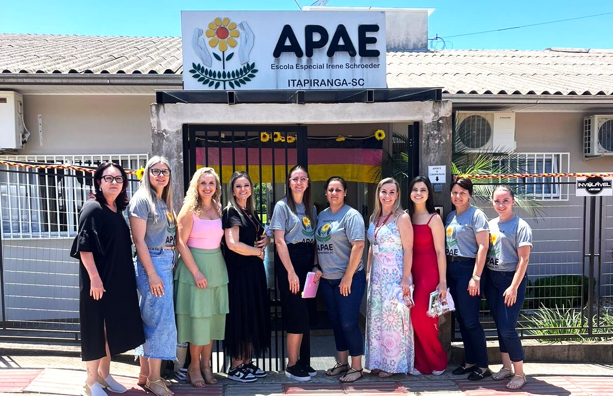 Um grupo de nove mulheres brancas estão em pé em frente ao prédio da Apae. Elas estão sorridentes. a primeira mulher é morena, cabelos pretos, vestido preto e usa óculos. A segunda é loira, usa óculos e está de blusa cinza e saia jeans. A terceira é loira, está de blusa rosa e saia verde. A quarta veste um vestido preto, cabelo castanho liso. A quinta tem cabelos pretos lisos, usa óculos e esta de blusa cinza e saia preta. A sexta mulher veste blusa cinza, usa óculos e está de calça jeans. A sétima  está com um vestido florido com predominância de azul e róseo. A oitava mulher tem o cabelo preto liso, com vestido vermelho. A nona mulher da imagem tem cabelo curto, camisa cinza e calça jeans. a última da foto está com cabelo preso e está de camisa cinza e calça jeans.