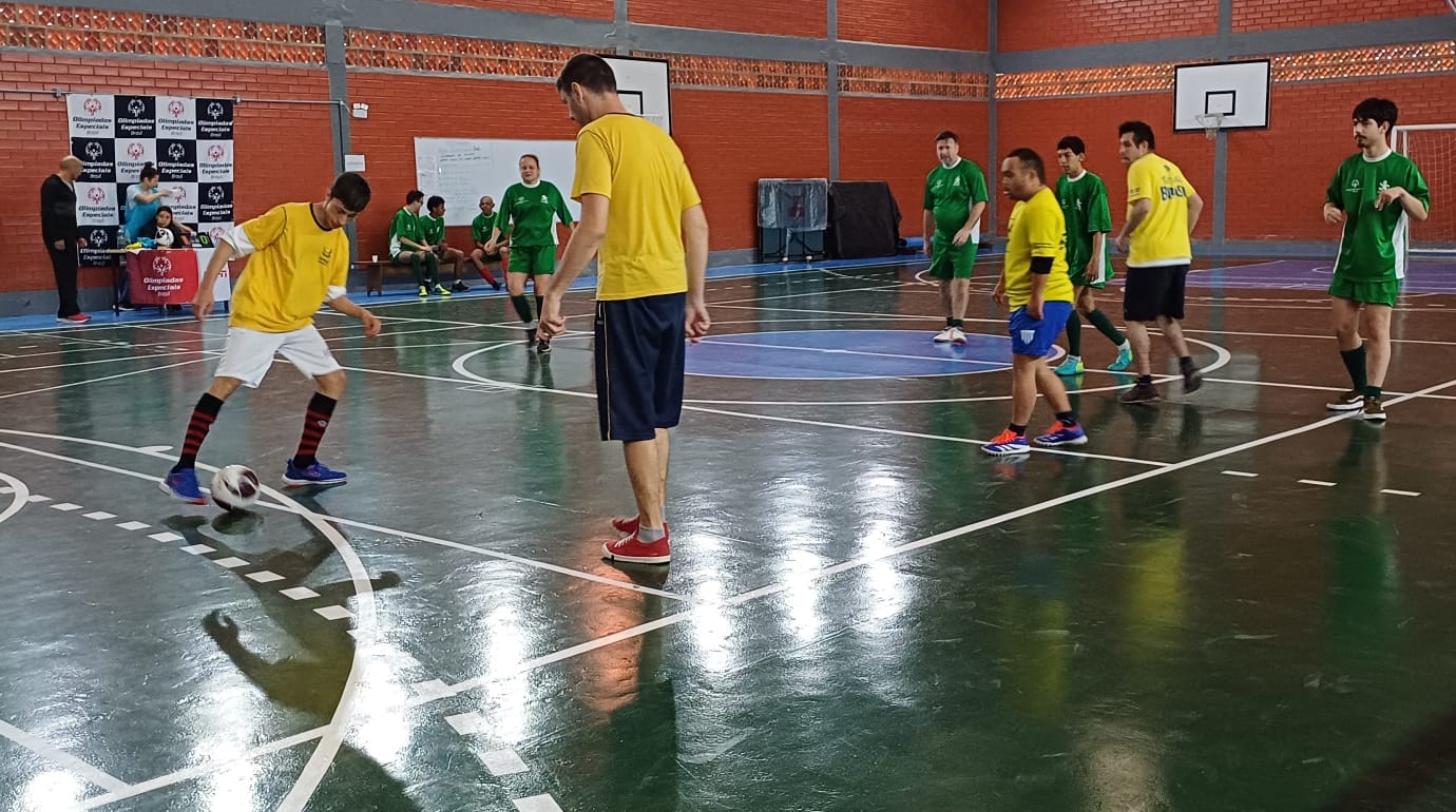 A imagem mostra uma partida de futsal em uma quadra esportiva. Na quadra central dois jogadores vestindo camisa amarela e verde disputam uma bola enquanto os demais jogadores, seis, observam a cena