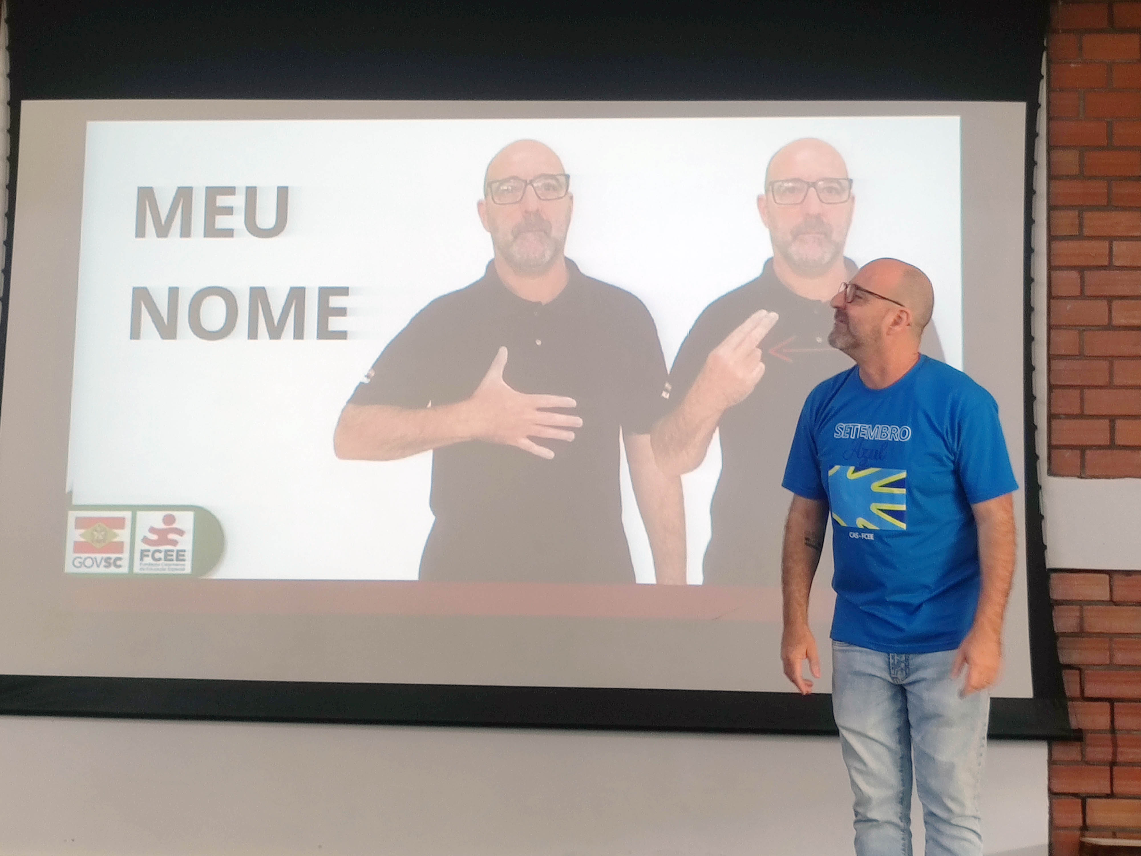 Homem vestido de azul em frente a telão onde aparece imagem dele fazendo sinal em Libras e o texto Meu Nome