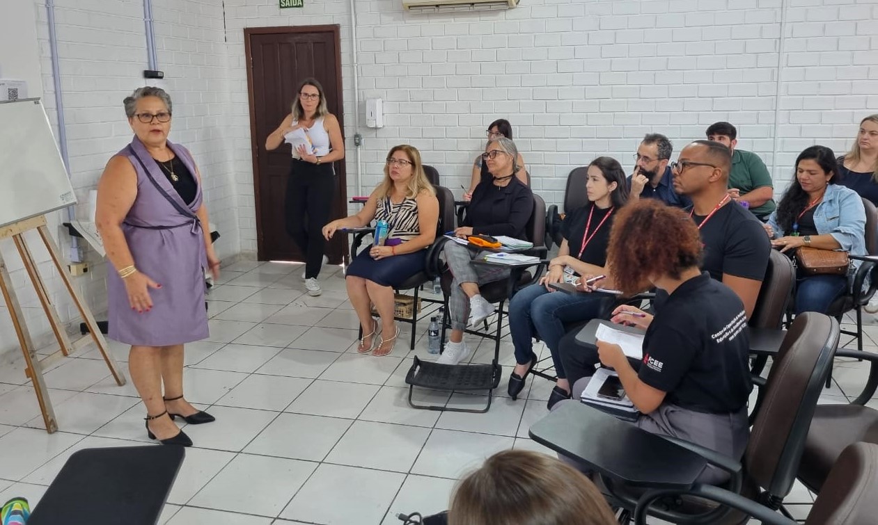 Uma mulher do lago esquerdo de uma sala está sendo observada por um grupo de pessoas em uma sala de aula. a mulher é branca e veste vestido lilás