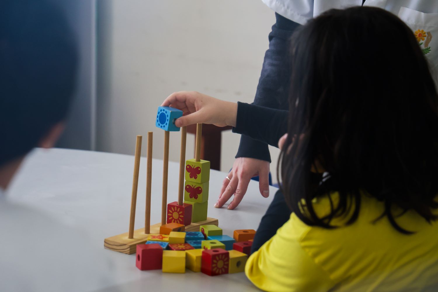 Imagem de uma menina de costa brincando com um brinquedo de madeira que está sobre uma mesa. A menina tem cabelos pretos e usa uma camiseta amarela