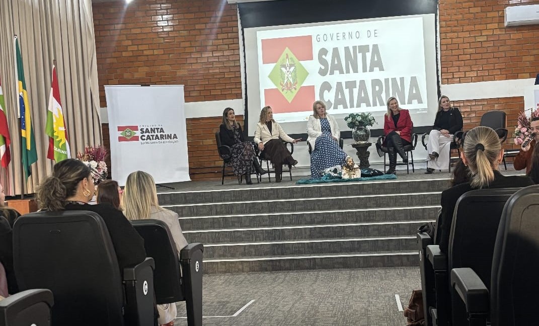 Cinco mulheres brancas estão sentadas em cadeiras em um palco em um auditório. Ao fundo tem a imagem da bandeira de Santa Catarina com os dizeres: Governo de "Santa Catarina". Em primeiro plano aparece três mulheres sentadas em poltronas. Elas são brancas e estão ouvindo as mulheres que estão no palco.