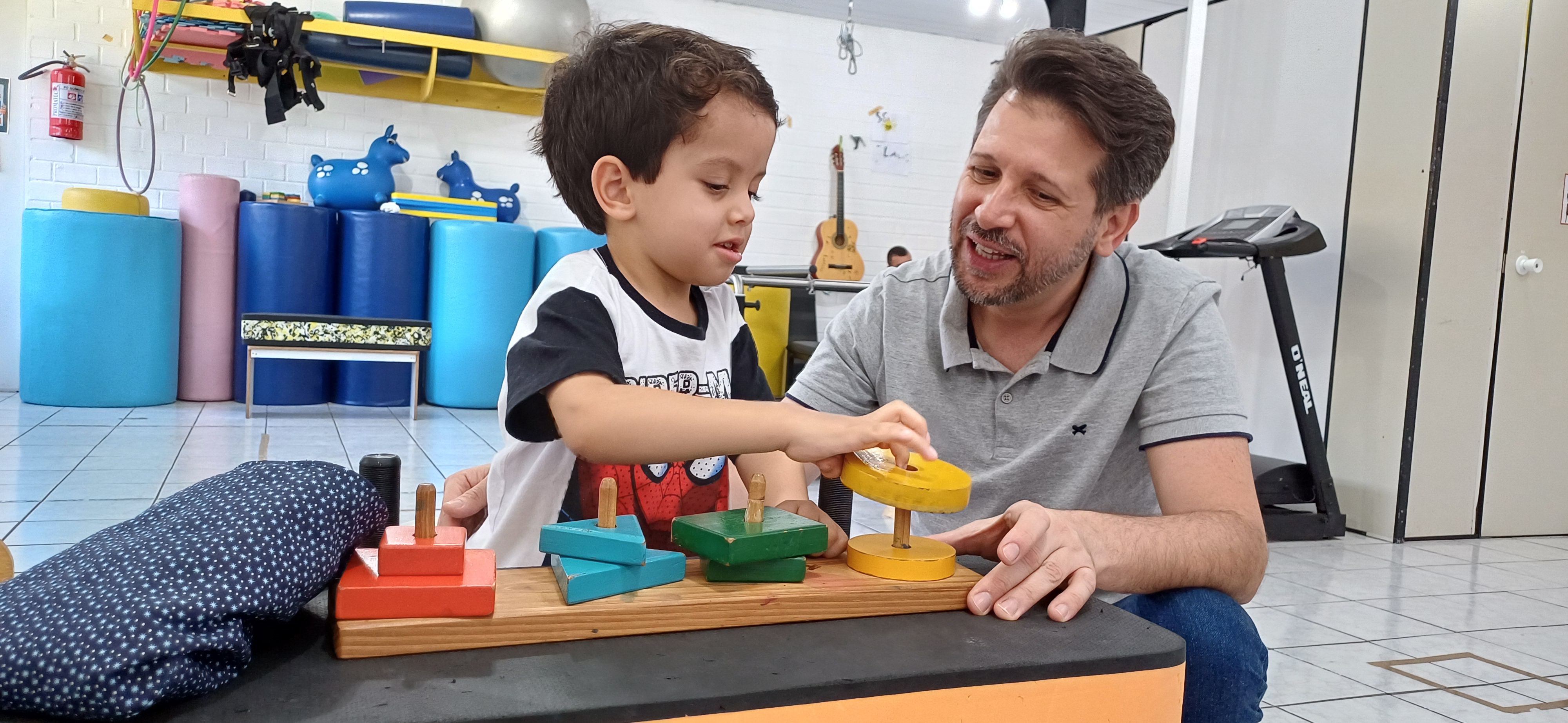 Professor com uma criança brincando sobre uma mesa com brinquedos lúdicos. O professor está sorrindo, ele é branco, cabelos pretos de camisa cinza. A criança é branca, cabelos pretos, camisa branca com gola preta. A criança interage com uma argola amarela e coloca sobre um pino de madeira. Sobre a mesa tem ainda formas de brinquedo em vermelho , azul e verde, que estão acopladas a pinos de madeira em forma de brinquedo. Ao fundo da imagem aparecem diversos colchoes azuis. A cena se passa em uma sala.