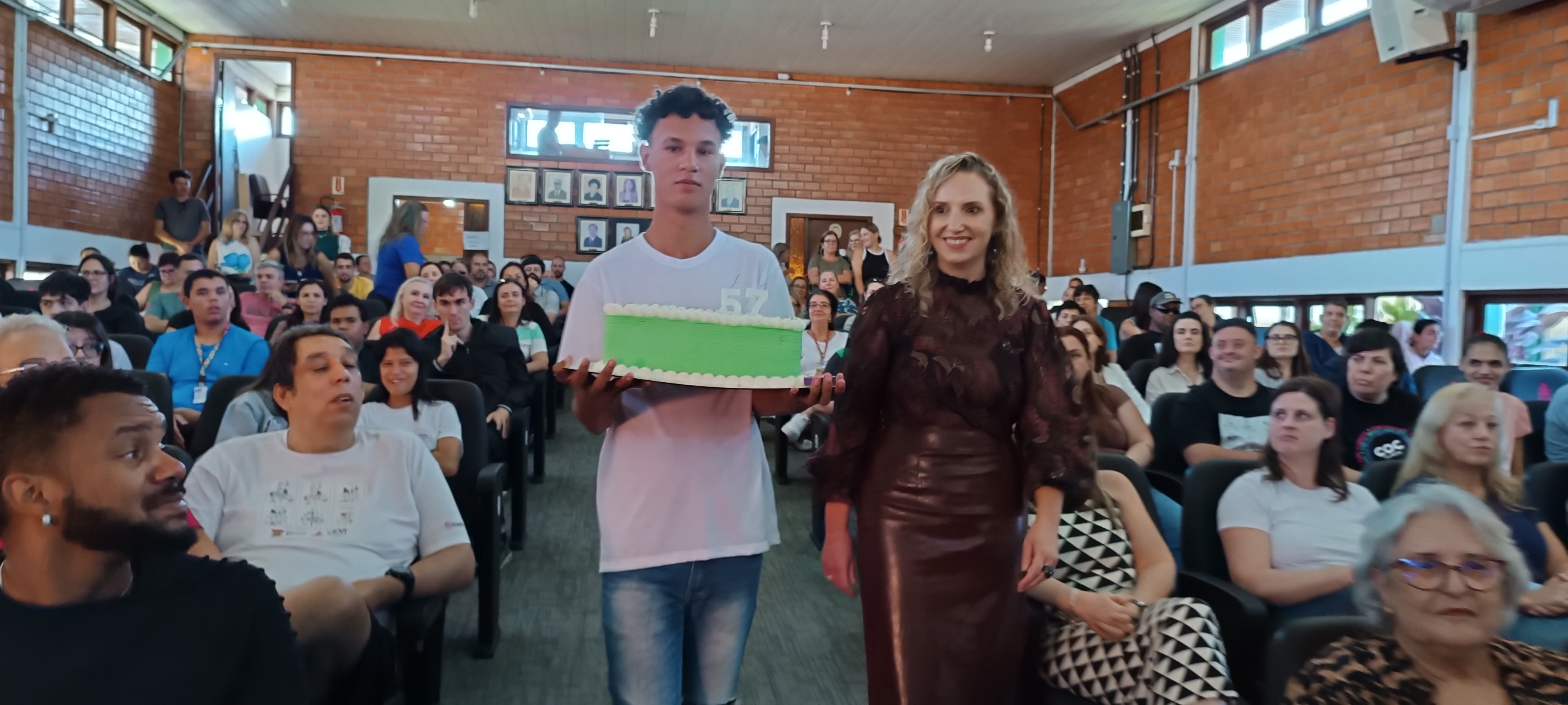 1 menino e 1 mulher caminham segurando bolo verde de 57 anos em meio ao auditório lotado