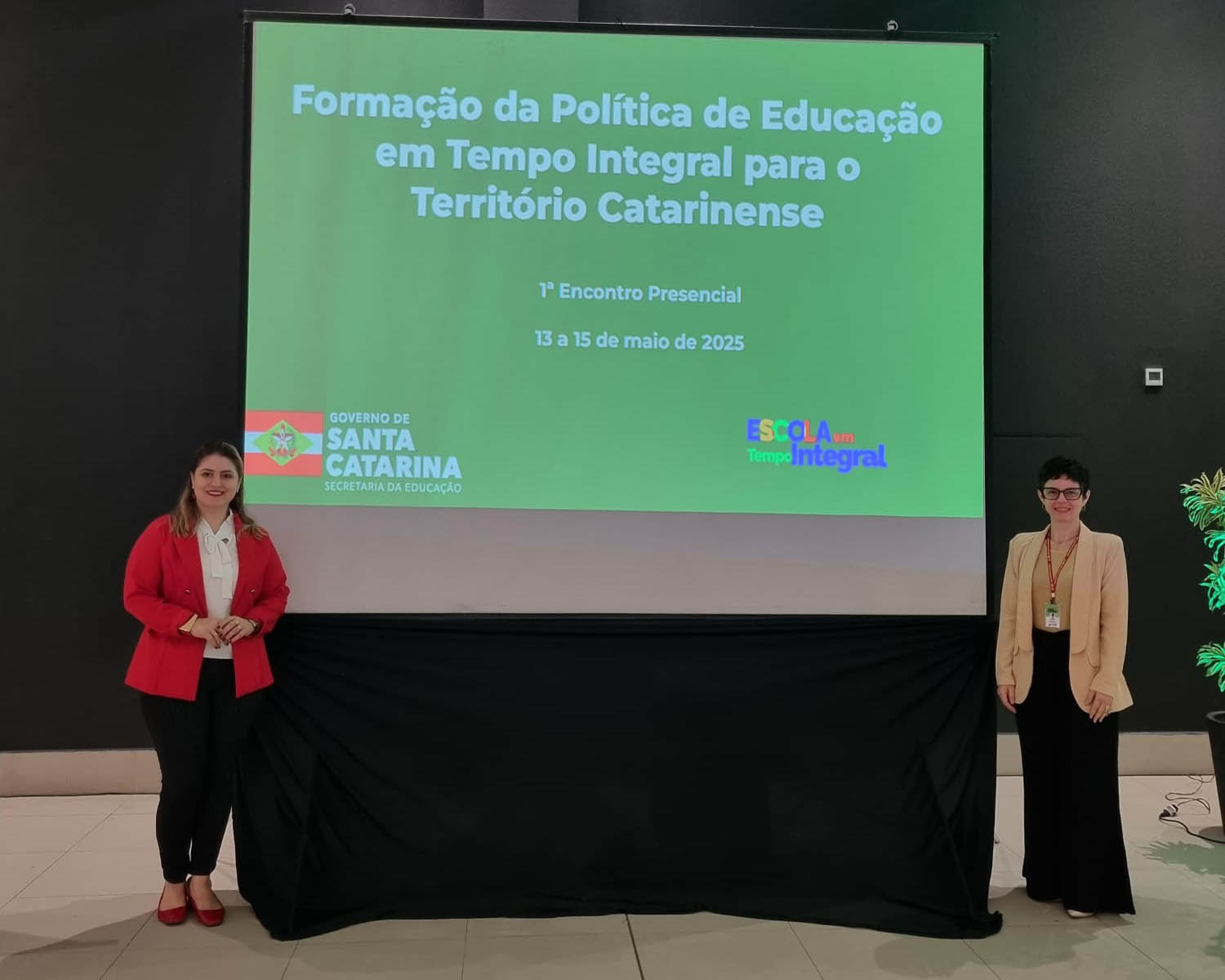 duas mulheres em pé posam para foto em placa escrito Formação da Política de Educação em Tempo Integral