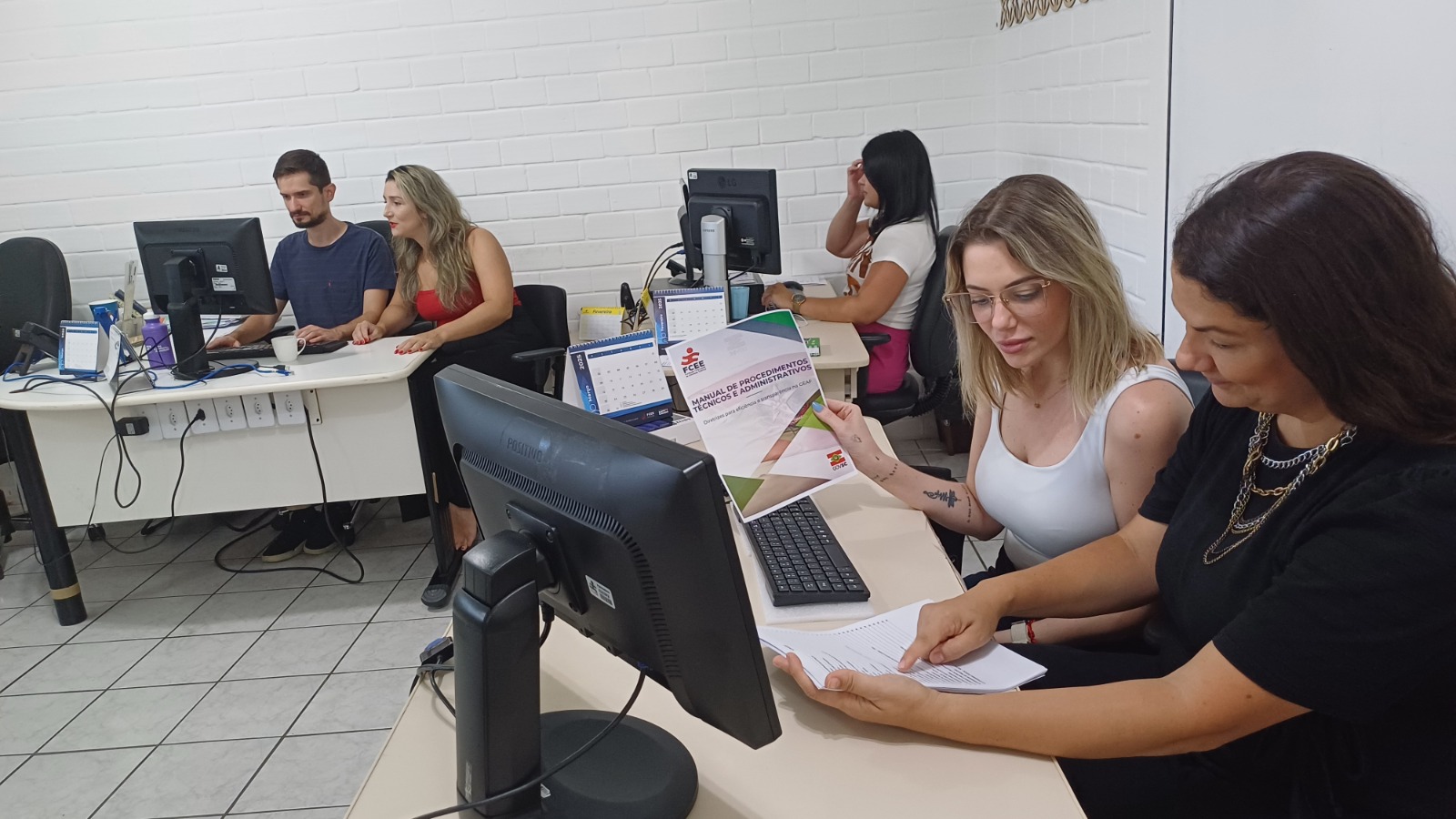 sete pessoas sentadas em mesas de trabalho, olhando para computador ou analisando papeis