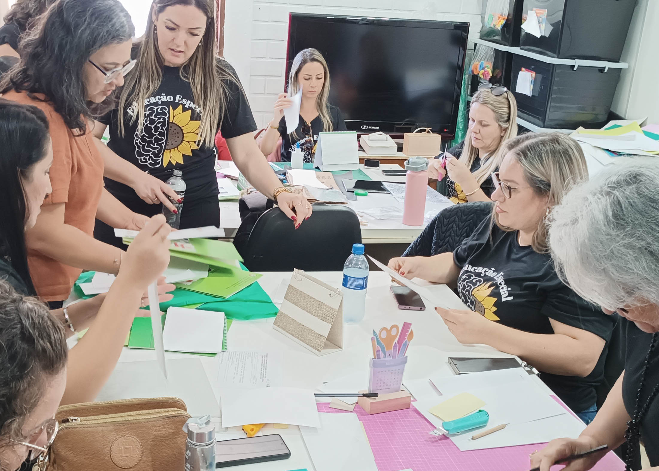 Sete mulheres em volta de mesa mexendo em materiais como papeis, tesouras.